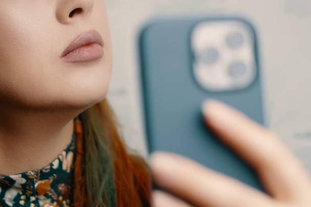 Rudowłosa dziewczyna z jasnym makijażem robi selfie na aparacie nowoczesnego telefonu. Blogerka ze smartfonem w ręku. Modne kobiece hobby. - Zdjęcie, obraz