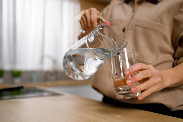 Vue rapprochée de la femme versant de l'eau de la cruche dans le verre dans la cuisine - Photo, image