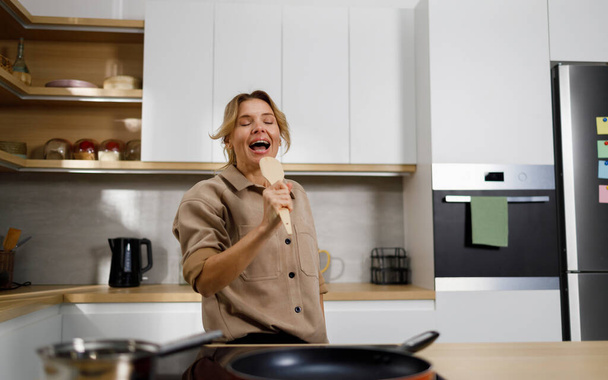 Ευτυχισμένη ενήλικη γυναίκα που τραγουδάει με μια ξύλινη σπάτουλα στην κουζίνα - Φωτογραφία, εικόνα