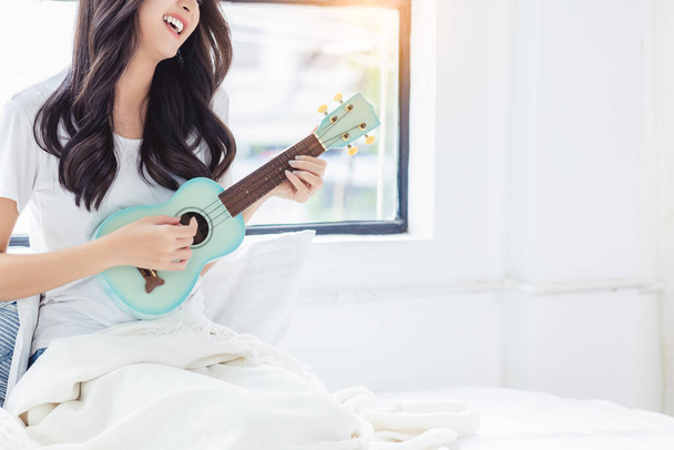Gyönyörű fiatal nő játszani kis gitár vagy ukulele gitár otthon ázsiai fiatal lány szeret játszani ukulele és énekelni egy dalt a boldogság és mosoly arc Ő tanul ukulele gitár Hobby szabadidő - Fotó, kép