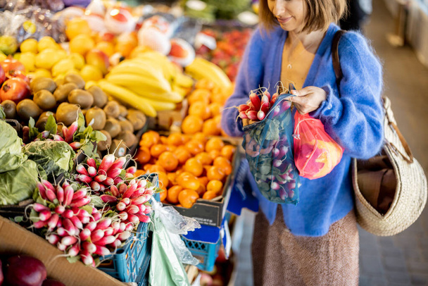 Woman takes a bunch of radishes from the counter, buying fresh vegetables and fruits at the local market. Shopping with reusable mesh bag. Sustainability and organic food concept - Photo, Image