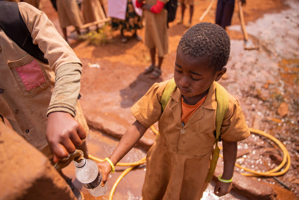 Pequeña colegiala africana recoge agua en la botella del grifo de la fuente ubicada en el patio de la escuela. - Foto, Imagen