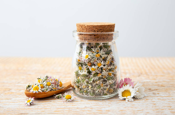 Sušená bylinná léčivá rostlina Common Daisy, známá také jako Bellis Perennis. Suché květy květin ve skleněné nádobě a dřevěné lžíci, připravené na přípravu bylinkového čaje, vnitřní zátiší. - Fotografie, Obrázek