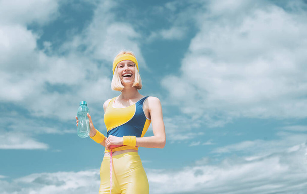 Cheerful smiling sporty girl with bottle of water at blue cloudy sky background. Beautiful blonde woman girl doing sport activities outdoor at fresh air. Sport is good for health - Φωτογραφία, εικόνα