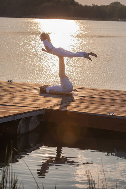 Güzel genç bir çift gün batımına karşı iskelede akro yoga yapıyor. Adam iskelede yatıyor ve kadını ayaklarıyla dengeliyor.. - Fotoğraf, Görsel