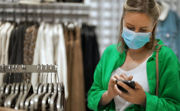 Vrouw met medisch masker die kleding kiest in de winkel. - Foto, afbeelding