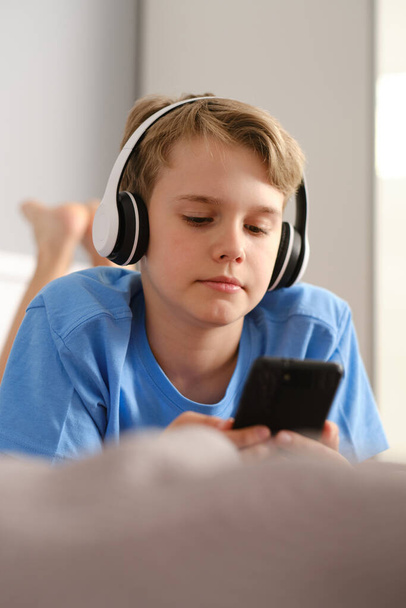 ヘッドフォンでかわいい男の子のクローズアップ肖像画。スマートフォンを持った10代の若者が家のベッドに横たわっている. - 写真・画像