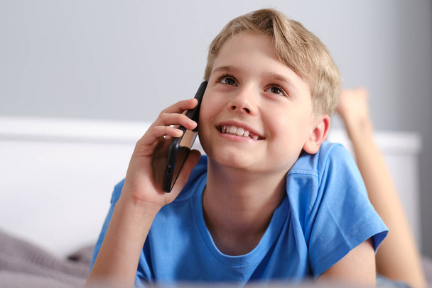 Portret uroczego chłopca rozmawiającego przez telefon leżącego na łóżku w domu. Bezprzewodowa w domu, rozmowa przez telefon. - Zdjęcie, obraz