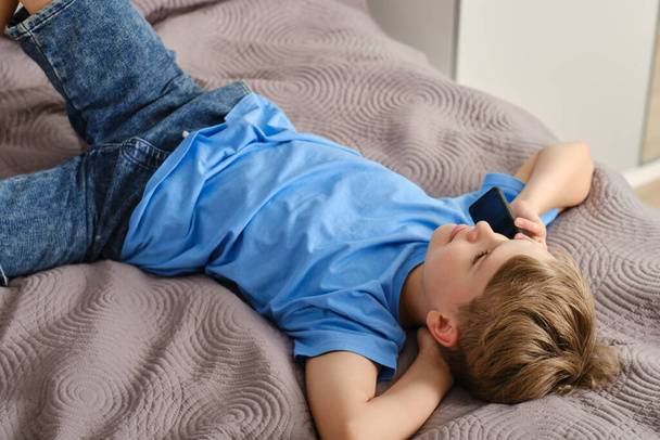 Χαλαρωμένο χαρούμενο αγόρι μιλάει στο τηλέφωνο ενώ είναι ξαπλωμένο στο κρεβάτι. Ασύρματο στο σπίτι, κινητή συνομιλία. - Φωτογραφία, εικόνα