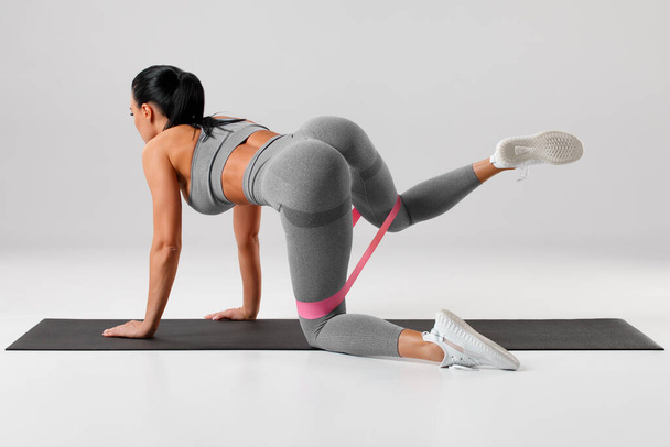 Sportovní dívka dělá cvičení pro hýždě s páskou odporu na šedém pozadí. Fitness žena cvičení ven - Fotografie, Obrázek