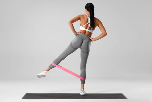 Athletic girl doing exercise for glutes with resistance band on gray background. Fitness woman working out - Fotoğraf, Görsel