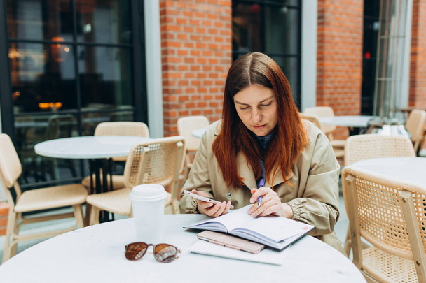 Mladá žena si vychutnává kávu, sedí na kavárně na terase v moderní městské ulici. Pracovnice pracující na mobilním telefonu stojící venku. Osoba sedící u stolu a zapisující si poznámky - Fotografie, Obrázek
