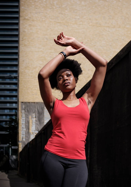 Jonge zwarte vrouw die zich uitstrekt in een straat in Londen. Ze draagt een rood mouwloos t-shirt. krullend haar. - Foto, afbeelding