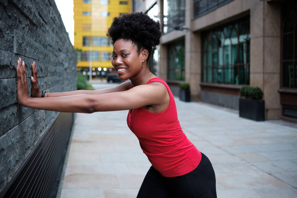 Portrait d'une jeune femme noire souriante s'étirant contre un mur. Elle est entre deux bâtiments et porte un t-shirt rouge sans manches. Cheveux bouclés. - Photo, image
