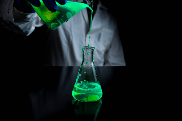 Eine Wissenschaftlerin experimentiert mit einer grün fluoreszierenden Lösung in einem Glaskolben in einem dunklen Chemielabor für die Entwicklung der Gesundheitsmedizin. Kopierraum schwarzer Hintergrund - Foto, Bild
