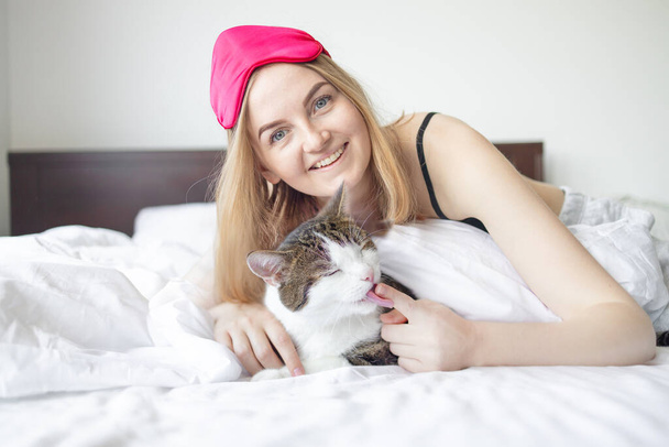 Νεαρή ξανθιά 30άρα γυναίκα με μάσκα ύπνου και χαριτωμένη γάτα ξαπλωμένη στο κρεβάτι στο δωμάτιο στο σπίτι.  - Φωτογραφία, εικόνα