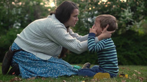 Το παιδί κλαίει στο πάρκο στην αγκαλιά της μαμάς. Η μαμά ηρεμεί το αγοράκι. Οικογενειακή μητέρα και παιδί με δάκρυα στα μάτια. συναισθηματικά. Αγαπώντας τη νεαρή μητέρα αγκαλιάζει και καταπραΰνει τον μικρό της γιο στο πάρκο. Μητρότητα. - Φωτογραφία, εικόνα