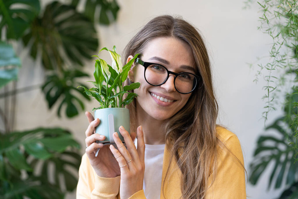 Portrait of beautiful florist woman in glasses looking at camera, holding pot with home plant near face in flower shop or greenhouse, blurred monstera on background. Hobby, houseplant lovers concept.  - Fotó, kép