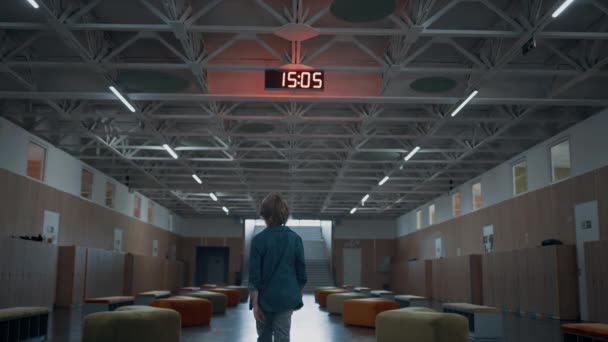Rear view schoolboy walking empty hall after classes. Modern elementary school interior with electronic clock on ceiling. Teen boy passing classroom lockers in corridor. Student wandering campus alone - Footage, Video