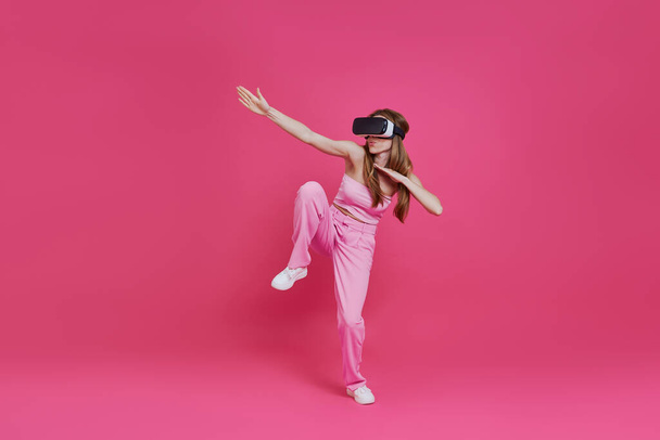 Полная длина игривая молодая женщина в гарнитуре виртуальной реальности весело на розовом фоне - Фото, изображение