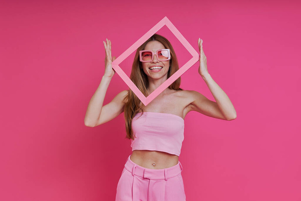 Aufgeregte junge Frau blickt durch einen Bilderrahmen, während sie vor rosa Hintergrund steht - Foto, Bild