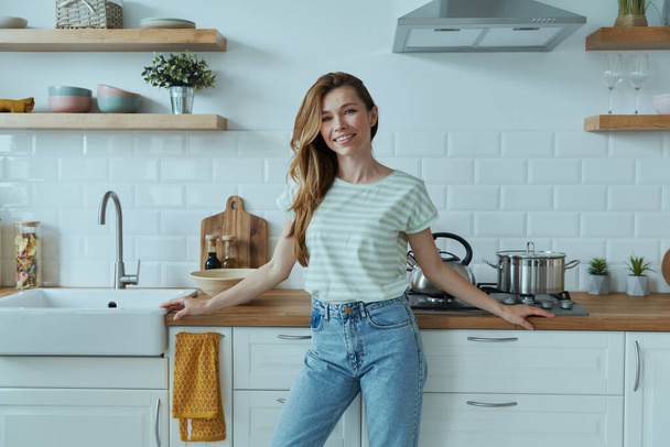 Beautiful young woman leaning at the kitchen desk and smiling - Photo, Image