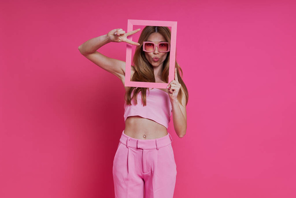 Playful young woman looking through a picture frame while standing against pink background - Photo, Image