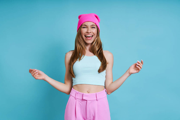 Boldog fiatal nő rózsaszín kalapban mosolyog, miközben áll ellen kék háttér - Fotó, kép