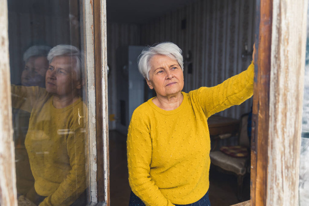 Ανώτερη Καυκάσια γυναίκα που κοιτάζει προσεκτικά έξω από το παράθυρο μεσαίου μεγέθους πυροβόλησε ηλικιωμένους σε εσωτερικούς χώρους και την ιδέα καραντίνας. Υψηλής ποιότητας φωτογραφία - Φωτογραφία, εικόνα