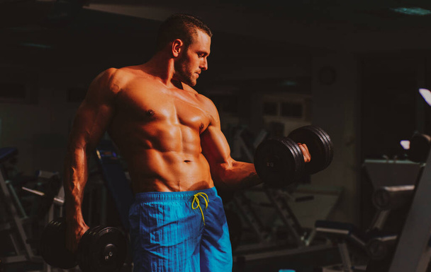 Shirtless strong bodybuilder training biceps in gym. Dumbbells exercises. Sportsman with shirtless torso. Sporty workout. Athletic body - Foto, afbeelding