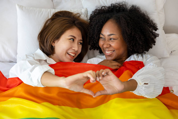 Para małżeństw tej samej płci z różnych ras z flagą tęczy LGBTQ dla dumy miesiąc leżąc razem na łóżku z gestem serca w celu promowania równości i różnic pojęć homoseksualnych - Zdjęcie, obraz