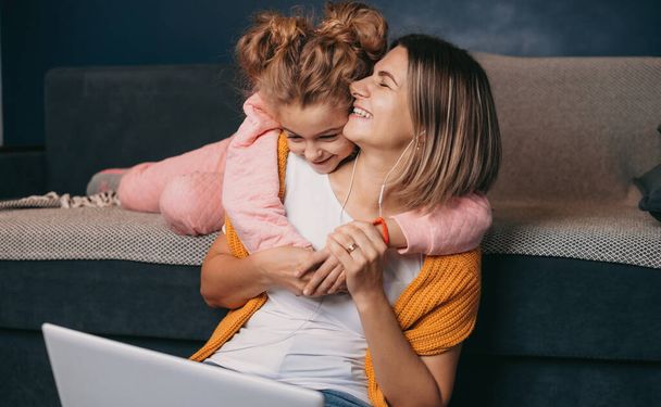 Freelancer woman working at home with laptop sitting on the floor near the sofa and behind her little daughter hugging mom around the neck. Mom work in modern - Photo, Image