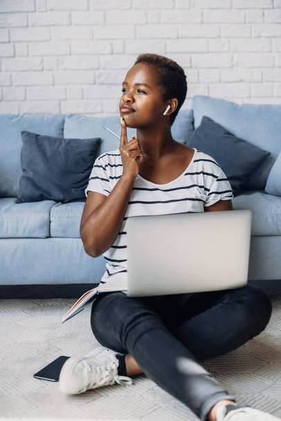 African american woman studying remotely at home using a computer laptop looking away thinking about question, sitting on the floor. Curly black hair. Pensive - 写真・画像