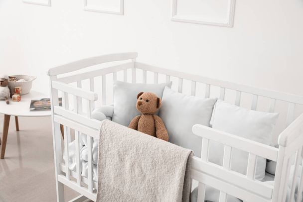 Baby crib with blanket, pillows and teddy bear near light wall - Foto, Bild