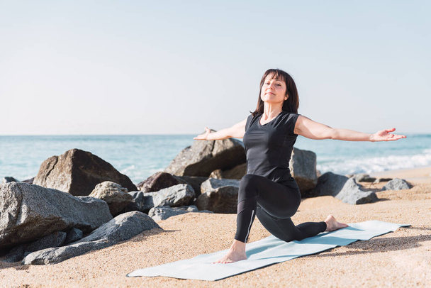 Corps complet de contenu femelle flexible debout à Anjaneyasana avec les bras tendus sur le tapis sur la plage de sable pendant la pratique du yoga en été - Photo, image