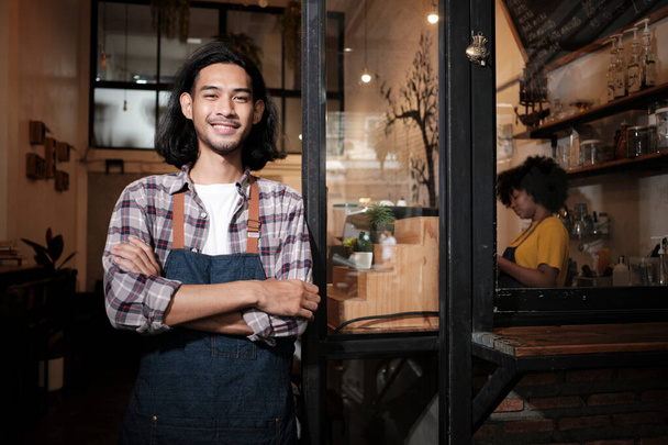 Un largo pelo guapo asiático hombre startup barista con delantal de pie en la puerta de la cafetería casual, los brazos cruzados, mirando a la cámara con una sonrisa acogedora, feliz y alegre con trabajos de servicio de cafetería. - Foto, Imagen