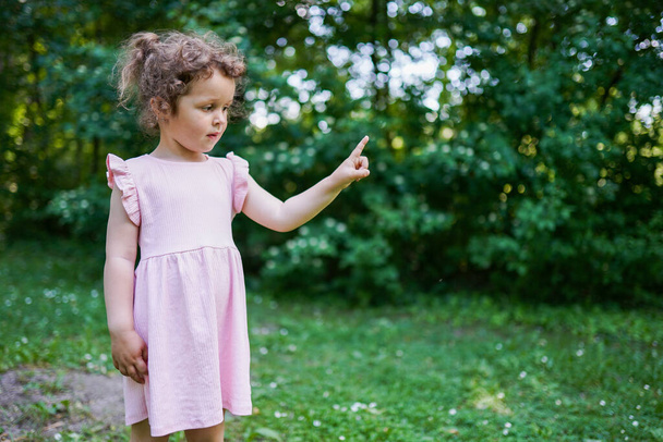 Niña en vestido rosa señala su dedo a un lado. Niña con pelo rizado en el parque. Moda y belleza de verano - Foto, imagen