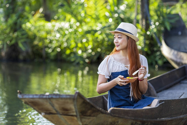 若いアジアの女性観光客は、タイの水上市場で木製のボートで旅行しており、東南アジア観光のための地元の通りの食べ物を持っています - 写真・画像