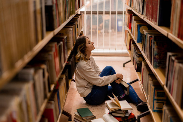Kütüphanede otururken kitaplığa yaslanan kız. - Fotoğraf, Görsel