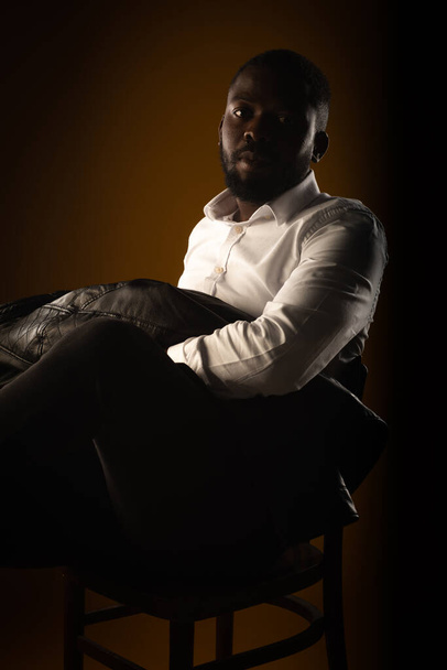 Siyah adamın portresi sandalyede poz verirken mükemmel bir duruş sergiliyor. - Fotoğraf, Görsel