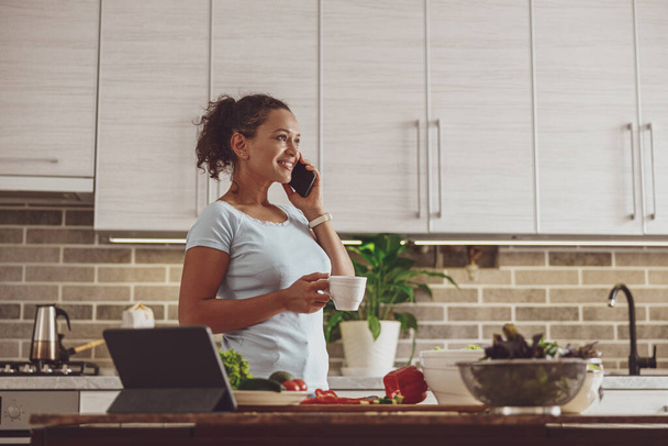 台所でお茶を飲みながら電話で話す女性サラダの野菜のスライスから気をそらして - 写真・画像