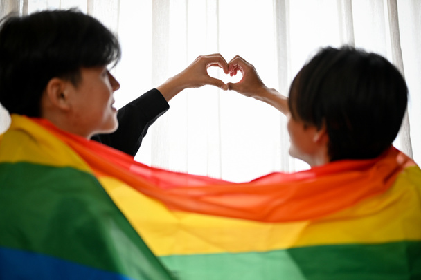 Szerető ázsiai meleg férfiak, akik szívformát öltenek kezükkel, szivárvány zászlóval borítva. LMBT meleg pasik. - Fotó, kép