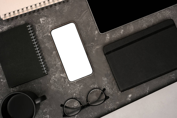 Σύγχρονη χώρο εργασίας με smartphone λευκή οθόνη mockup, γυαλιά, βιβλίο, σημειωματάριο, σπιράλ σημειωματάριο και ένα φλιτζάνι καφέ σε γκρι τσιμέντο υφή φόντο. πάνω όψη, επίπεδο lay - Φωτογραφία, εικόνα