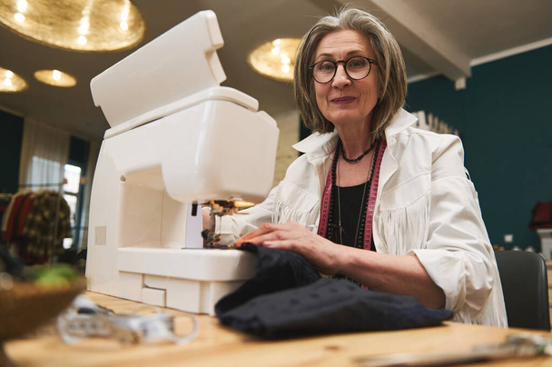 Elderly European woman dressmaker, seamstress, tailor or sewer in workshop studio, works on sewing machine, designing new collection clothes. Fashion designer business start-up, entrepreneur concept - Valokuva, kuva