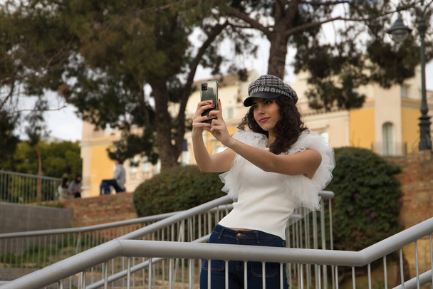 Young and beautiful woman, brunette, with curly hair, wearing white shirt, jeans and cap, taking pictures of herself with her cell phone. Concept beauty, fashion, model, trend, selfie, photos. - Photo, image