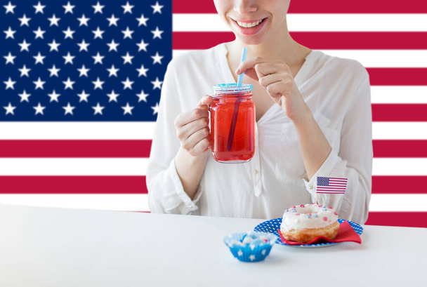 independence day, patriotism and holidays concept - close up of happy woman with iced donut drinking juice from mason jar glass at 4th july party over flag of united states of america on background - Photo, image