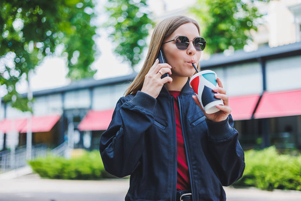 Menina bonita e jovem em óculos de sol escreve uma mensagem em um telefone celular e beber café ao ar livre
 - Foto, Imagem