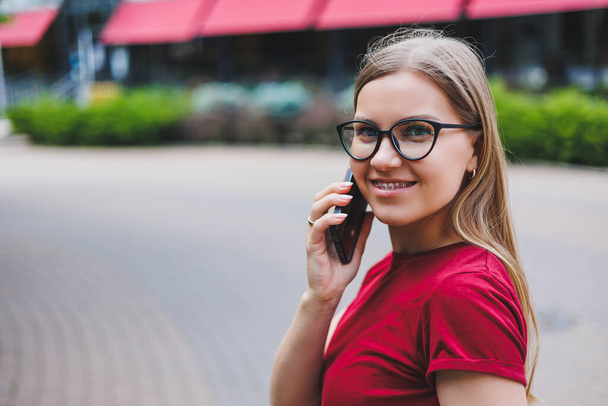 Young cheerful female in grey pullover and blue jeans and glasses standing on sidewalk carrying shopping bags and messaging on smartphone in New York - Fotoğraf, Görsel