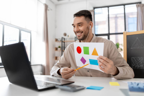 distance education, primary school and remote job concept - happy smiling male teacher with laptop and picture of geometric shapes in different colors having online class at home office - Foto, Imagen