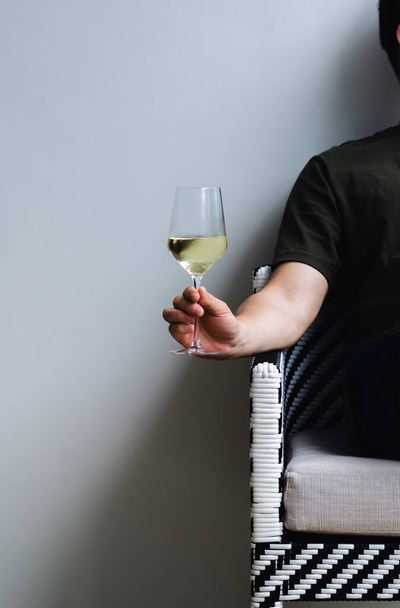 Ein Glas Weißwein in der Hand einer Person, die auf einem Stuhl mit Schatten auf weißem Wandhintergrund sitzt. - Foto, Bild